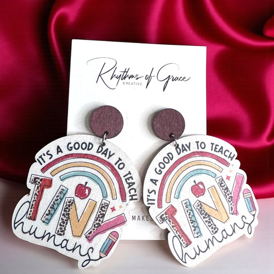 Earrings with rainbow design and text on a white card with 'Rhythms of Grace' branding, against a red satin background.