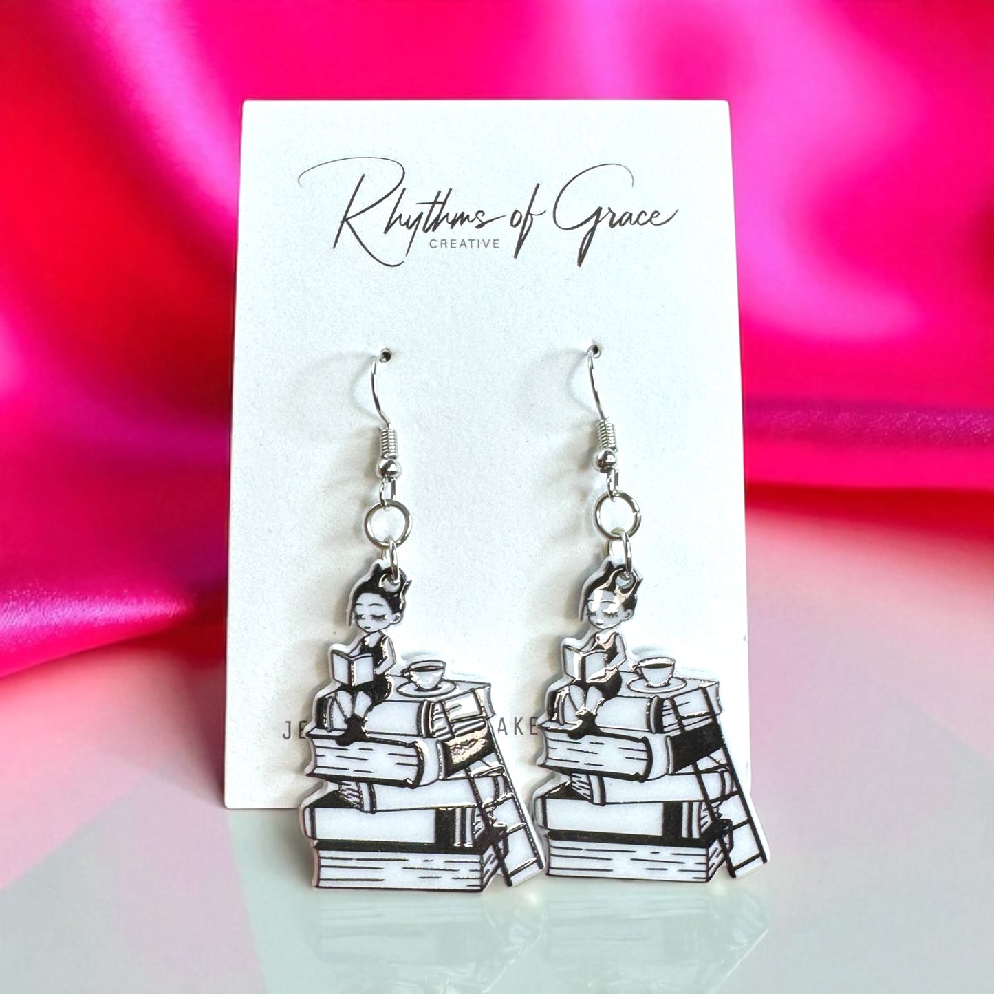 Earrings with a design of a person on books on a 'Rhythms of Grace' card with a pink and green background.