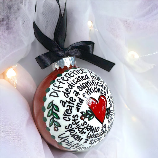 A hand-painted, round acrylic Christmas ornament with a green and red design, featuring a heart and leaves, and text that says 'Difference Maker'. It is adorned with a black ribbon at the top and has a white fabric background with fairy lights.