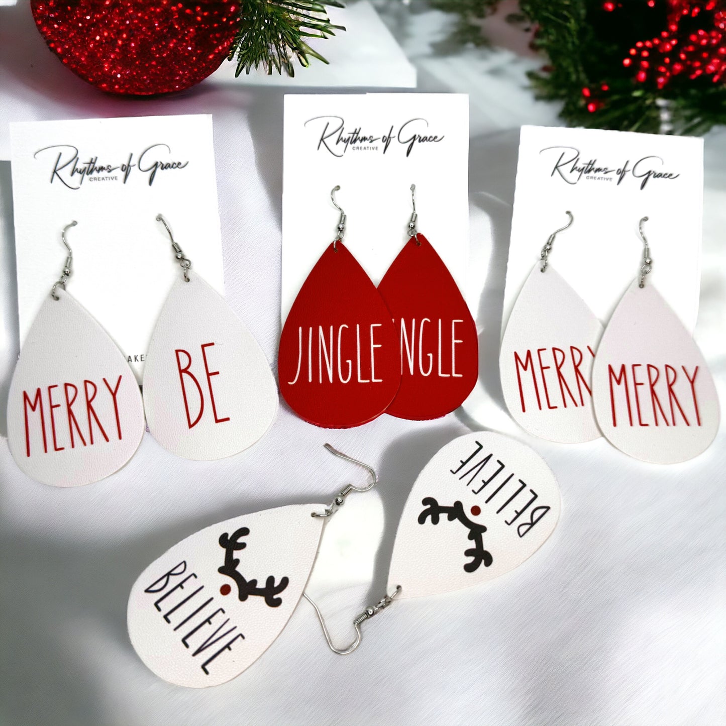 A set of Christmas-themed earrings with various designs, including a red reindeer, a white Christmas tree, and text phrases such as 'MERRY BE' and 'JINGLE JANGLE', displayed on a white card with a red and green Christmas tree in the background.