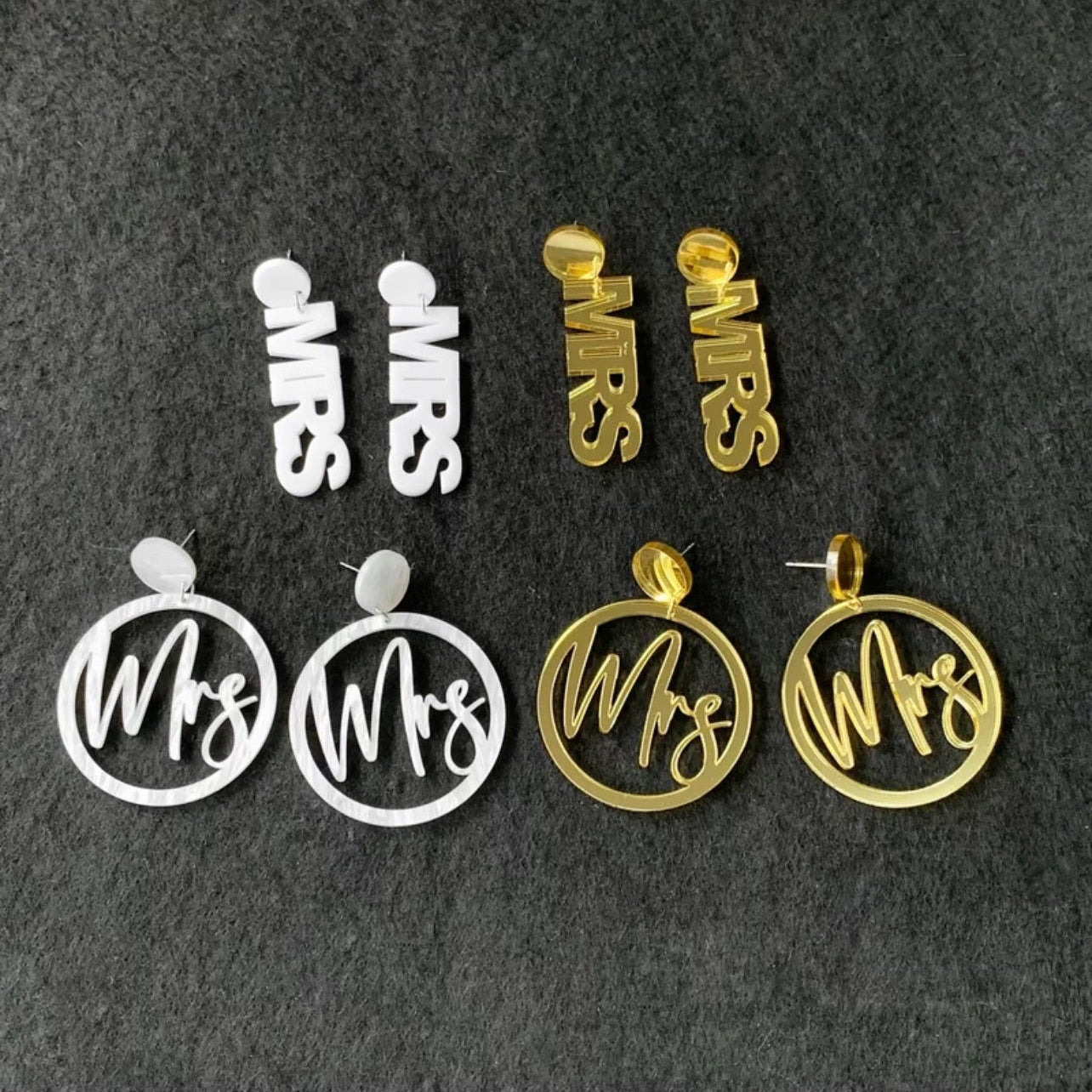 A pair of acrylic MRS earrings in gold and silver colors, with the letters 'MRS' engraved on them, displayed against a grey background.