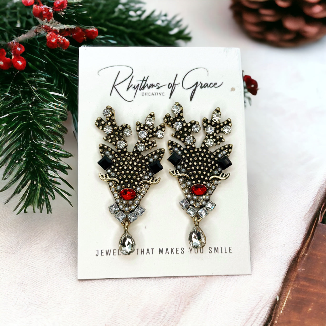 A pair of blue Christmas-themed reindeer earrings, with rhinestone embellishments, displayed on a card with 'Rhythms of Grace' branding.