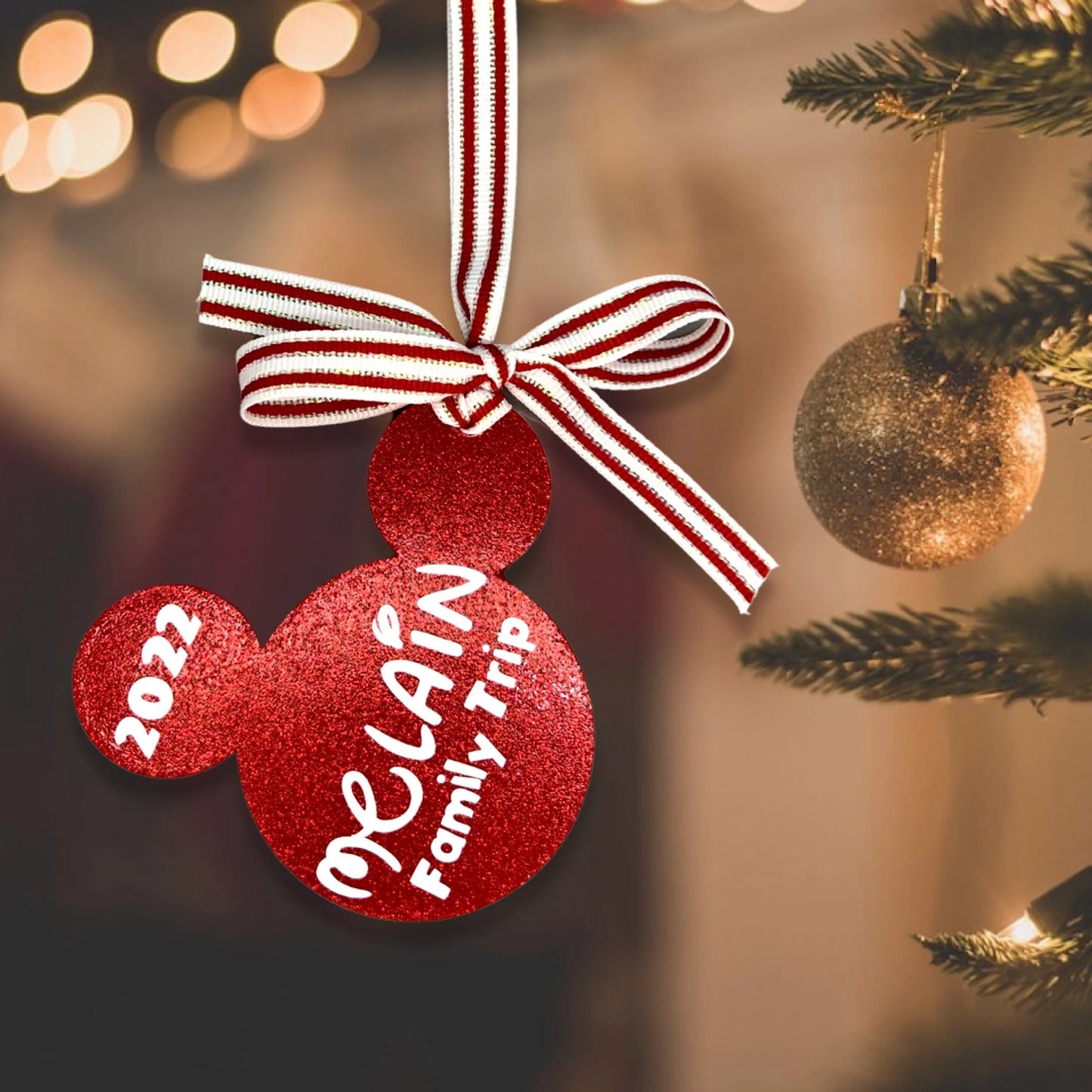 A personalized red glitter mouse ears ornament with '2024 Family Trip' printed on it, hanging from a white and red striped ribbon on a Christmas tree.