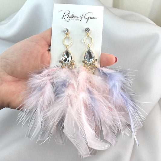 Feather earrings held by a hand with a 'Rhythms of Grace' card in the background.