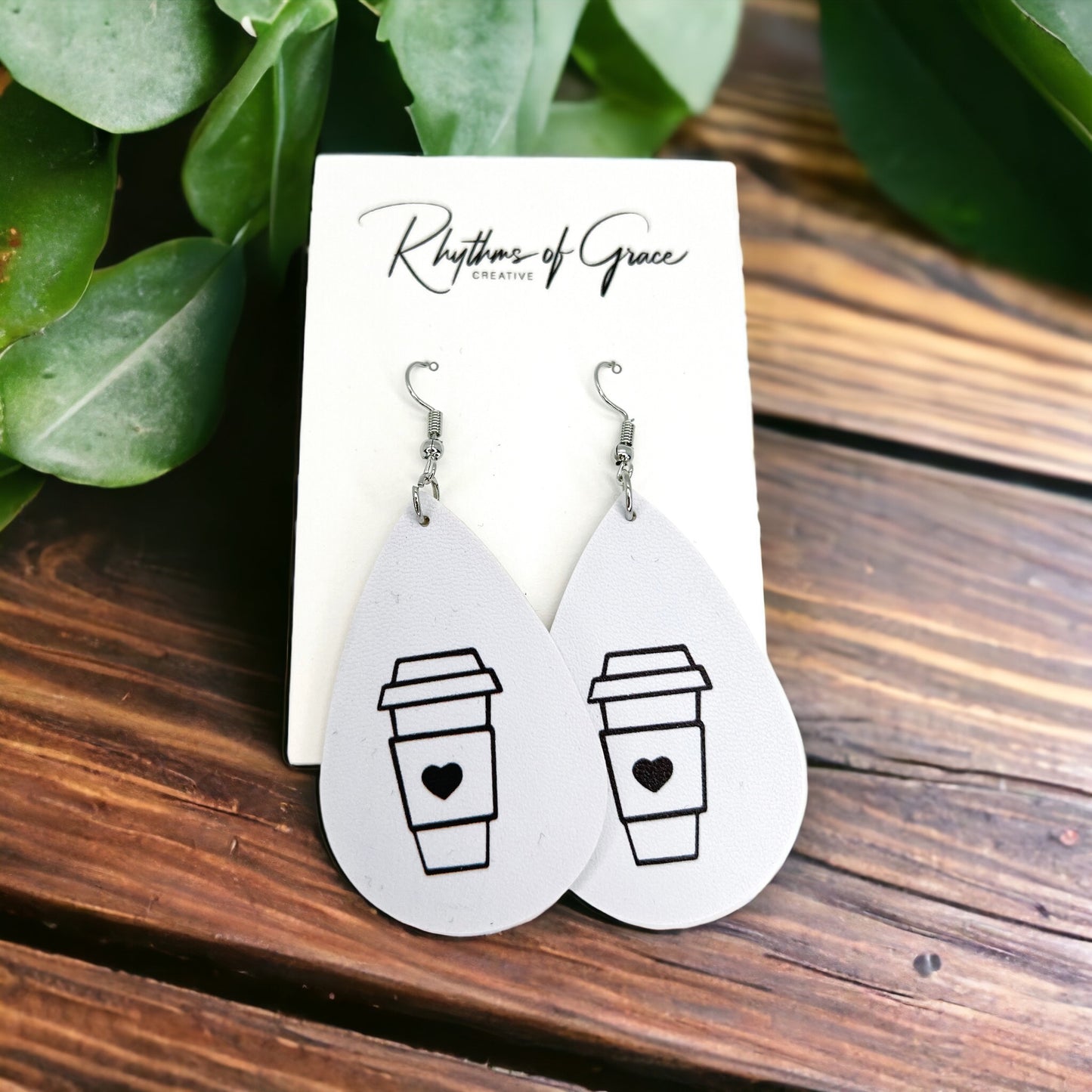 A pair of teardrop-shaped earrings with a coffee cup design, attached to a cardboard display with 'Rhythms of Grace' written on it, placed on a wooden surface with foliage in the background.