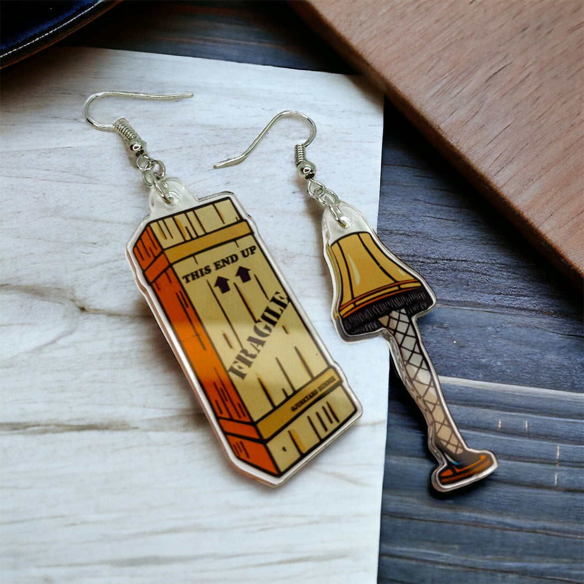A pair of earrings designed like a leg lamp from the movie 'Christmas Story', with the lamp shade in orange and the base in brown color, displayed on a wooden surface.