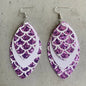 Three pairs of mermaid tail-shaped earrings displayed on cards, with the ocean in the background. Each earring features a unique color pattern with white, gold, purple, and green elements.