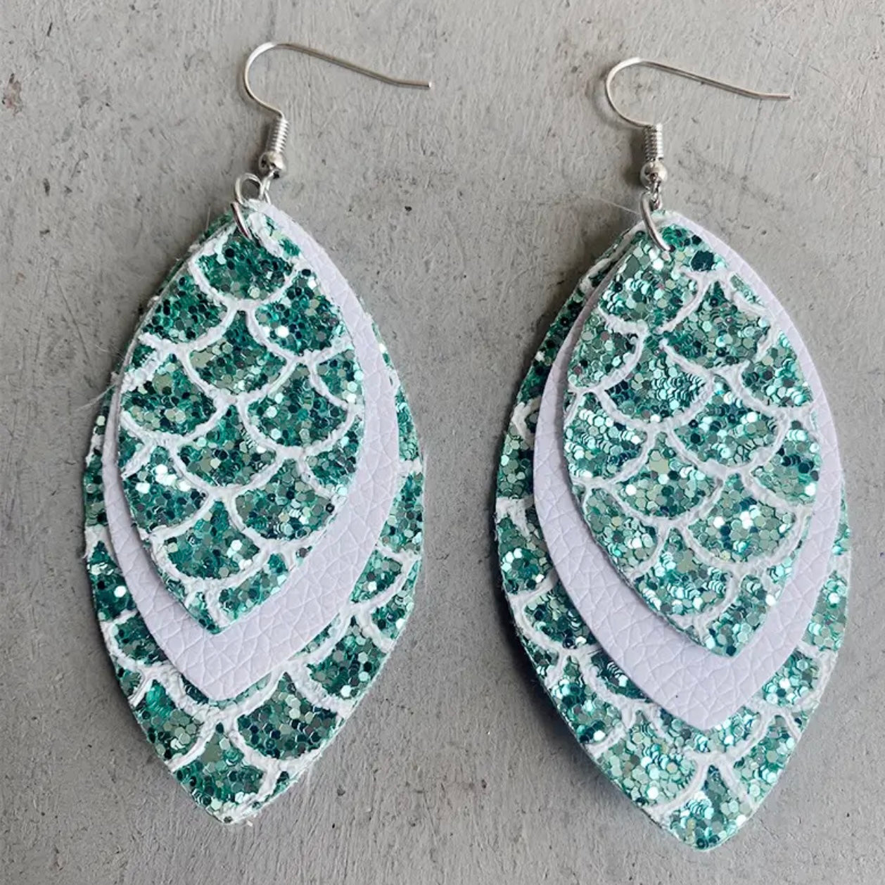 Three pairs of mermaid tail-shaped earrings displayed on cards, with the ocean in the background. Each earring features a unique color pattern with white, gold, purple, and green elements.