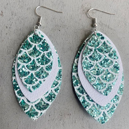 Three pairs of mermaid tail-shaped earrings displayed on cards, with the ocean in the background. Each earring features a unique color pattern with white, gold, purple, and green elements.