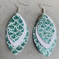 Three pairs of mermaid tail-shaped earrings displayed on cards, with the ocean in the background. Each earring features a unique color pattern with white, gold, purple, and green elements.
