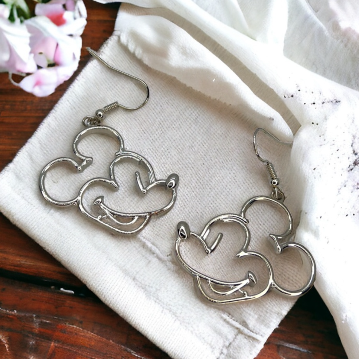 Pair of silver Mickey Mouse earrings on a white cloth with a wooden surface underneath.