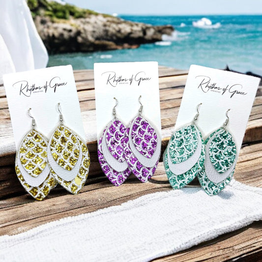 Three pairs of mermaid tail-shaped earrings displayed on cards, with the ocean in the background. Each earring features a unique color pattern with white, gold, purple, and green elements.