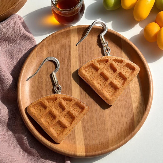 Waffle-shaped earrings on a wooden plate with a jar of honey and fruits in the background.
