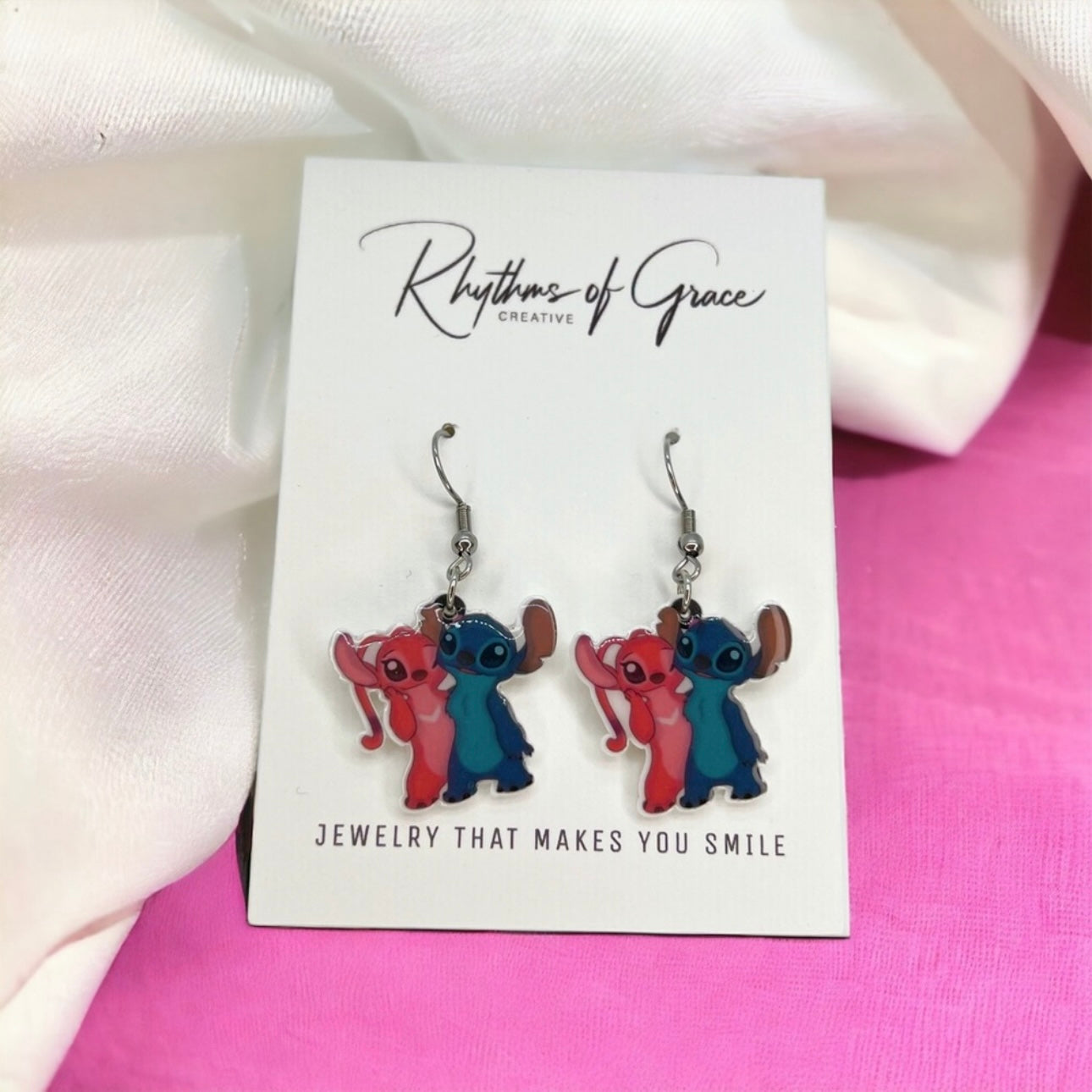 Pair of cartoon character earrings on a 'Rhythms of Grace' card with a pink and white background.
