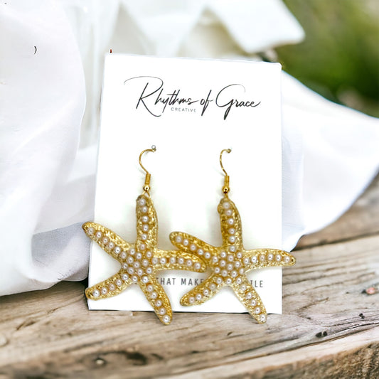 A pair of gold-colored starfish earrings adorned with faux pearls, displayed on a white card with 'Rhythms of Grace' written at the top.