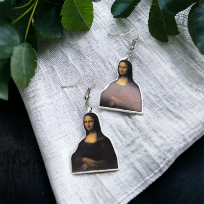 A pair of acrylic earrings featuring an image of the Mona Lisa, displayed on a white fabric surface with green leaves in the background.