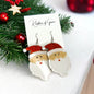 A pair of silver-colored Santa Claus earrings with red rhinestone details, displayed on a card with 'Rhythms of Grace' branding, set against a Christmas-themed backdrop.