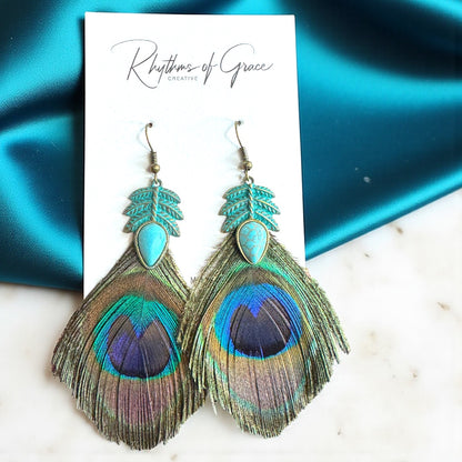 Peacock feather earrings on a 'Rhythms of Grace' branded card with teal fabric background