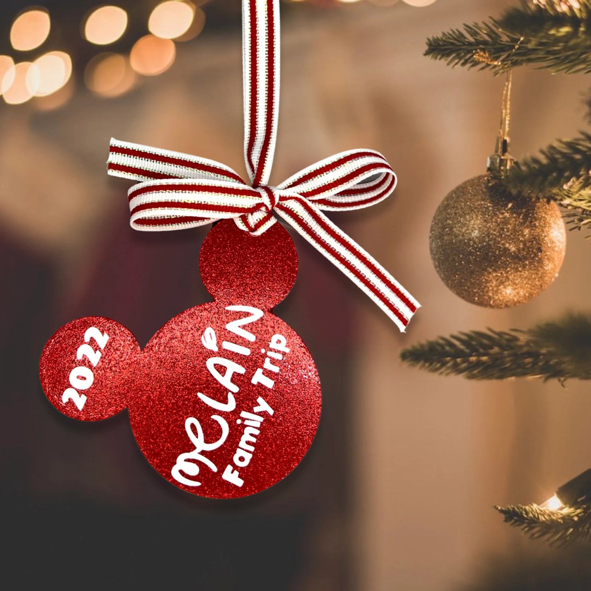 A personalized red glitter mouse ears ornament with '2024 Family Trip' printed on it, hanging from a white and red striped ribbon on a Christmas tree.