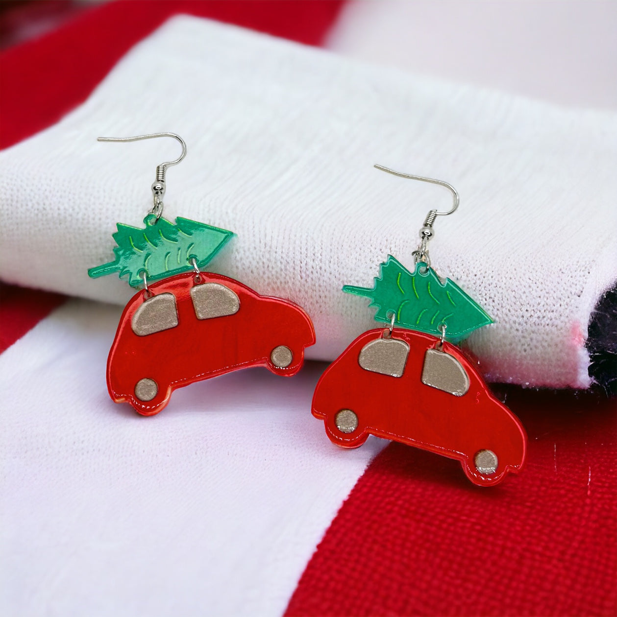 A pair of red Christmas truck earrings with green Christmas tree toppers, placed on a red and white fabric.