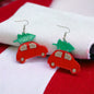 A pair of red Christmas truck earrings with green Christmas tree toppers, placed on a red and white fabric.
