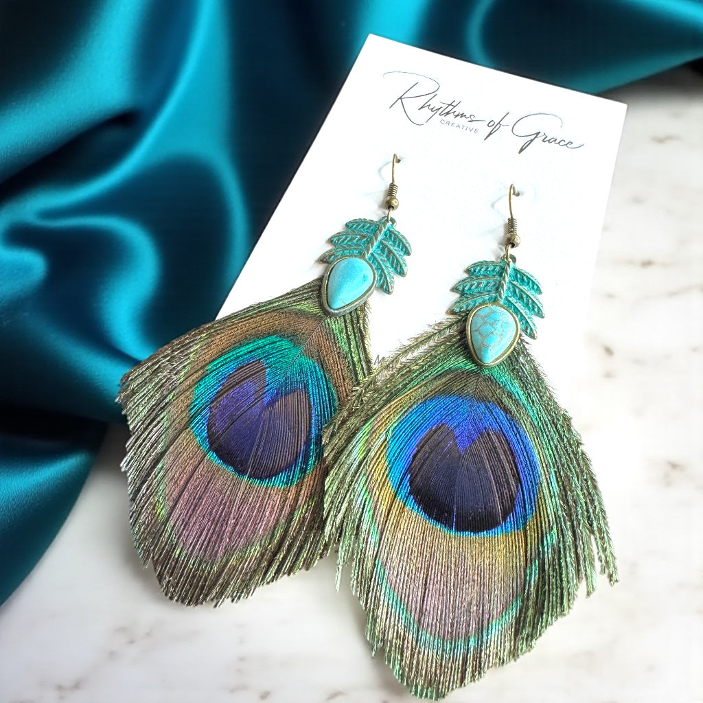 Peacock feather earrings on a white card with 'Rhythm of Grace' branding, against a blue satin background.